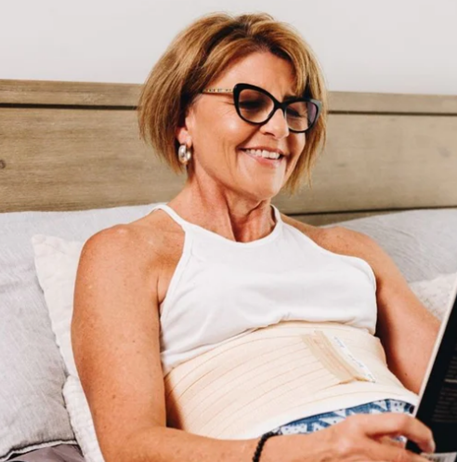 Smiling woman sitting up in bed reading, wearing a Hernia Belt in colour bisque.